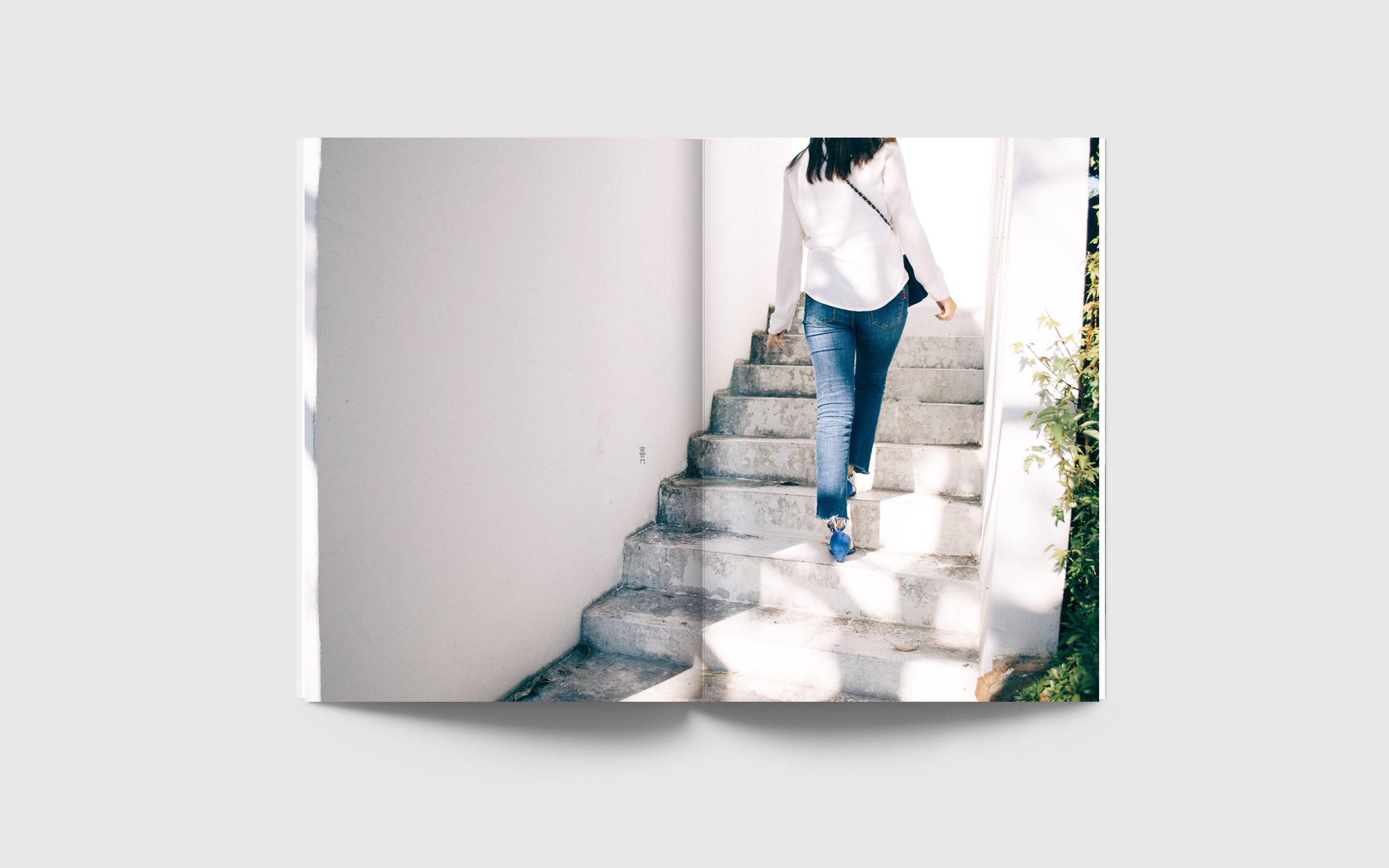 Spread: a full-bleed photograph of a woman in jeans and blue sandals climbing sunlit white steps, with a small vertical caption on the white wall